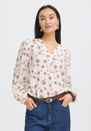 Kvinde med langt mørkt hår iført en hvid bluse med brune blomstertryk, blå jeans, et brunt bælte og guldøreringe, hænderne i lommerne.