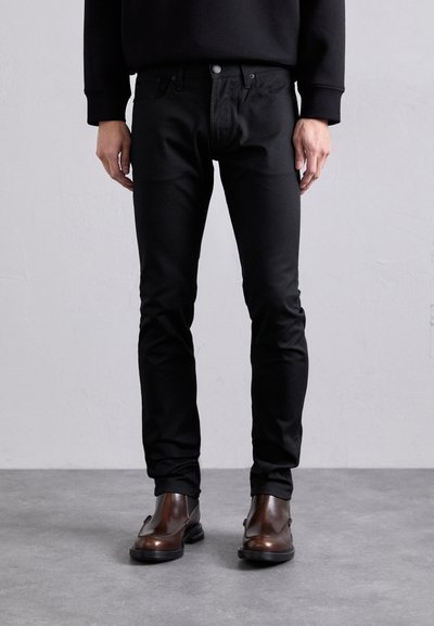 Černé slim-fit džíny s pěti kapsami, vyrobené z hladkého materiálu. V kombinaci s tmavě hnědými kotníkovými botami, které ukazují elegantní, moderní vzhled.