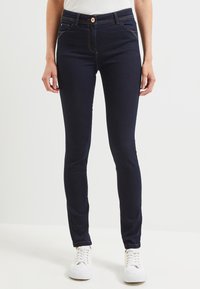 Jeans skinny in denim blu navy con una texture liscia, caratterizzati da tasche frontali e cuciture a contrasto. Indossati con sneakers bianche.