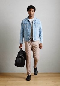 Veste en denim bleu clair sur un t-shirt blanc, pantalon beige et chaussures noires, tenant un sac de sport noir, sur un sol en bois.