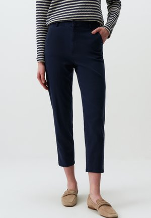 Navyblaue Chinos mit geradem Schnitt, ausgestattet mit einem Knopfverschluss, seitlichen Taschen und leicht verkürzter Länge. Kombiniert mit braunen Slippern.