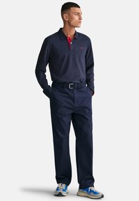 Tengerészkék, hosszú ujjú polo ing piros gallérral, sötét nadrággal és szürke, fekete akcentusú kék sportcipővel párosítva.