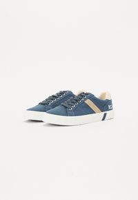 Blue canvas sneakers with beige stripe. White rubber sole and eyelets. Round toe design, lace-up front, and logo detail on the side.