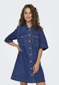 Denim shirt dress in dark blue, short sleeves, button front, two chest pockets, and light stitching details. Smooth texture, knee-length cut.