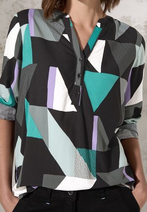 Femme portant une blouse multicolore à motif géométrique avec les manches retroussées et un pantalon noir, debout les mains dans les poches.