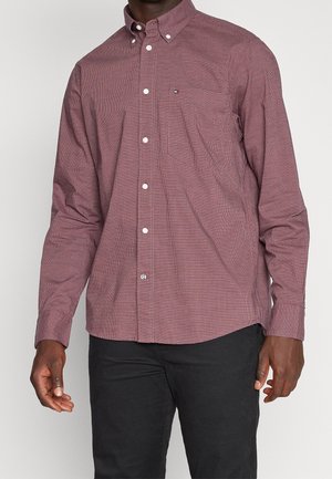 Homme portant une chemise à manches longues, boutonnée, à carreaux bordeaux, associée à un pantalon noir, debout devant un fond uni.