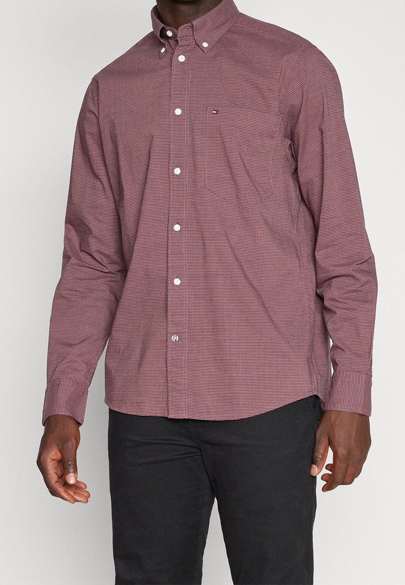 Homme portant une chemise à manches longues, boutonnée, à carreaux bordeaux, associée à un pantalon noir, debout devant un fond uni.