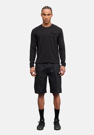 Uomo con capelli intrecciati in piedi, indossa una maglia nera a maniche lunghe con tasca, pantaloncini neri, calzini neri e scarpe da ginnastica nere su uno sfondo grigio.