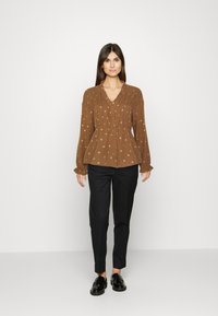 Blusa longa castanha de manga longa com decote em V, detalhes franzidos e bordado floral dourado, combinada com calças pretas slim fit e mocassins pretos.