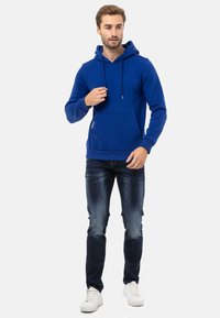 Blauer Hoodie mit Kängurutasche, Kordelzugkapuze und gerippten Bündchen, kombiniert mit verwaschenen blauen Jeans und weißen Turnschuhen.
