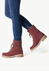 Botas de tobillo de ante burdeos con suelas de goma, ojales dorados y cordones. Llevadas con jeans ajustados azules, mostrando un diseño funcional y durabilidad.