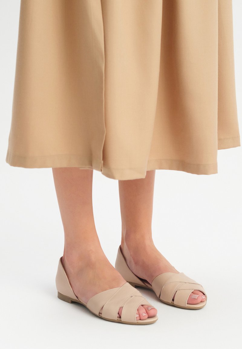 Beżowe sandały z otwartym noskiem, z krzyżującymi się paskami i niską, płaską podeszwą. Materiał wydaje się gładki z matowym wykończeniem. Idealne do codziennego użytku.