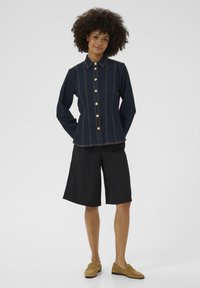 Džínová bunda s límečkem, zlatými knoflíky a kontrastním prošíváním, v kombinaci s černými šortkami a hnědými mokasíny. Bílý pozadí.