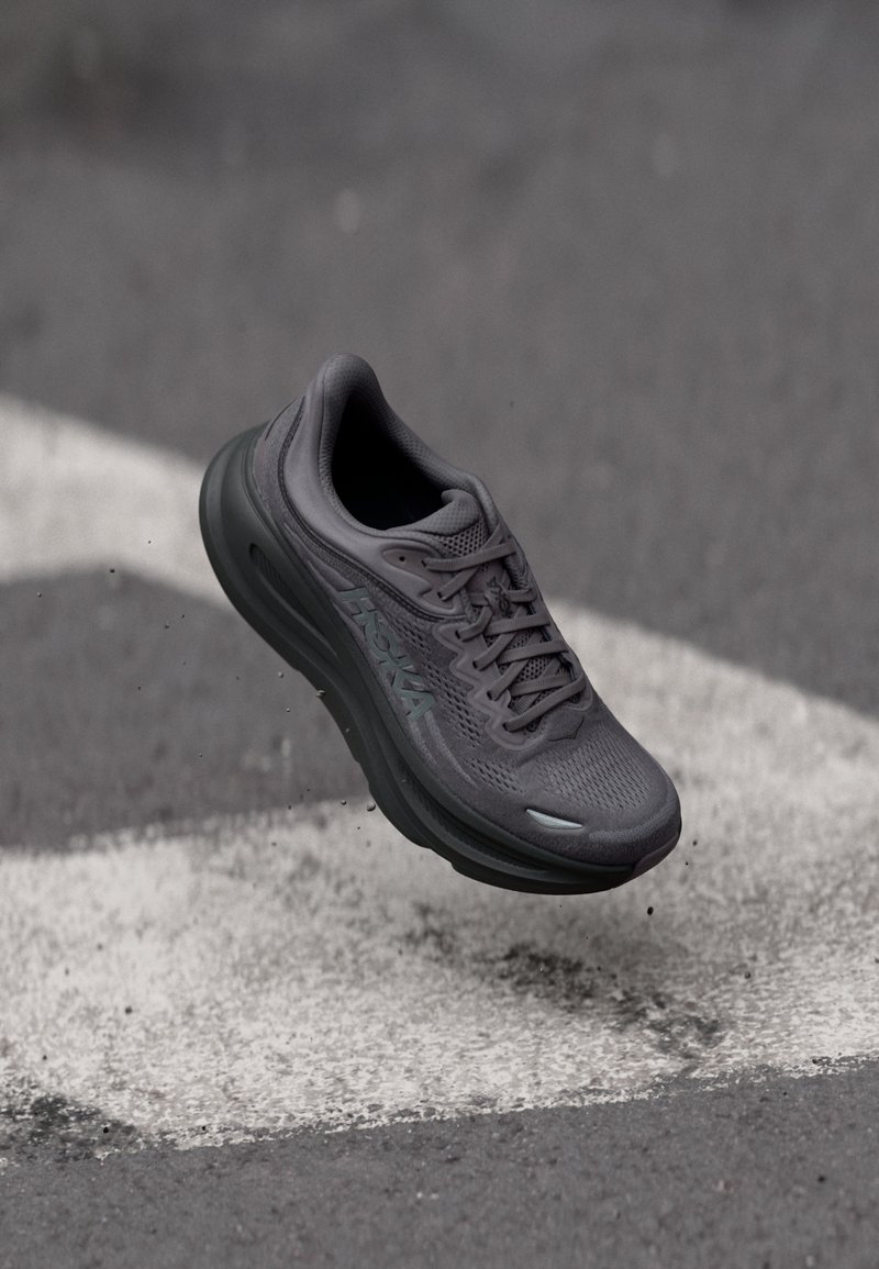 Γκρι αθλητικό παπούτσι Hoka αιωρείται στον αέρα πάνω από άσφαλτο με λευκές γραμμές δρόμου και μικρά σκουπίδια.