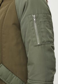 Olivovo zelená bomber bunda s prešívaným dizajnom, kontrastnými rukávmi a zipsovým vreckom so striebornými kovovými doplnkami na ľavom ramene.