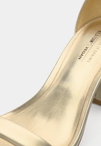 Χρυσό vegan δερμάτινο σανδάλι με ψηλό τακούνι, λεία υφή, σχέδιο με ανοιχτά δάχτυλα και λουράκι στον αστράγαλο· διαθέτει διακριτικό λογότυπο στην εσωτερική σόλα.