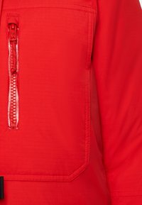 Giacca rossa in tessuto testurizzato, dotata di una tasca con zip e cuciture rinforzate. Il colore è uniforme, senza motivi o accenti visibili.