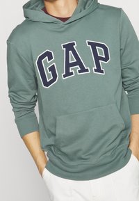Felpa verde con cappuccio realizzata in tessuto misto cotone, caratterizzata da un grande logo "GAP" blu navy, una tasca a marsupio e polsini aderenti.