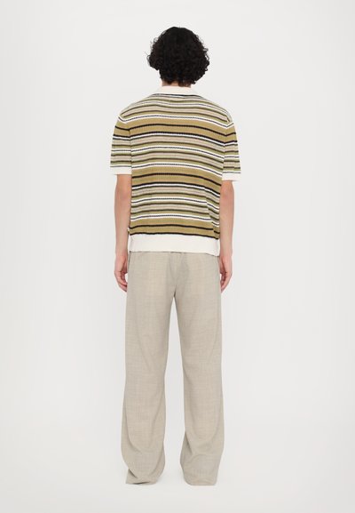 Maglietta a righe di maglia verde, beige e nera, con maniche corte, colletto a coste e parte posteriore leggermente più lunga. Abbinata a pantaloni beige chiaro.