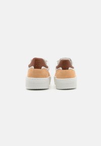 Weiße Sneaker mit tanfarbenen Wildlederakzenten an der Ferse, ausgestattet mit einem markierten Lederpatch und dicken weißen Gummi-Sohlen. Einfaches, sauberes Design.