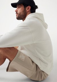 Cream hoodie with a textured finish, paired with beige shorts featuring a logo patch. The outfit is casual and relaxed in fit.