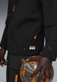 Sort atletisk jakke med orange detaljer og logo, udstyret med lynlåslommer og en glat tekstur, holdt ved siden af en marmor basketball.