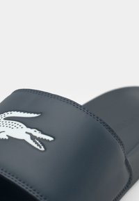 Navy blue slide sandal with a wide strap, featuring a white crocodile logo at the center and visible stitching along the edges.