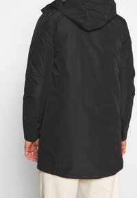 Veste noire isolante avec capuche, dotée d'une texture lisse et d'un dos plus long. Comprend des poches latérales et un design élégant.