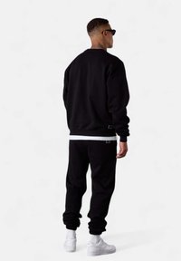 Ensemble de sweat-shirt et de sweat-pants noirs, avec des poignets côtelés, une doublure intérieure blanche et un détail de logo, associé à des baskets blanches.