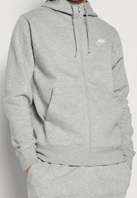 Felpa Nike grigia chiara con zip, tasche frontali e cappuccio con cordino, indossata con pantaloni della tuta grigi chiari abbinati.