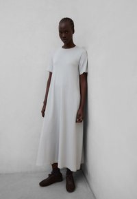 Femeie cu părul împletit, purtând o rochie lejeră, cu mâneci scurte, până la gleznă, de culoare gri deschis și pantofi sport maro închis, stând sprijinită de un perete simplu.