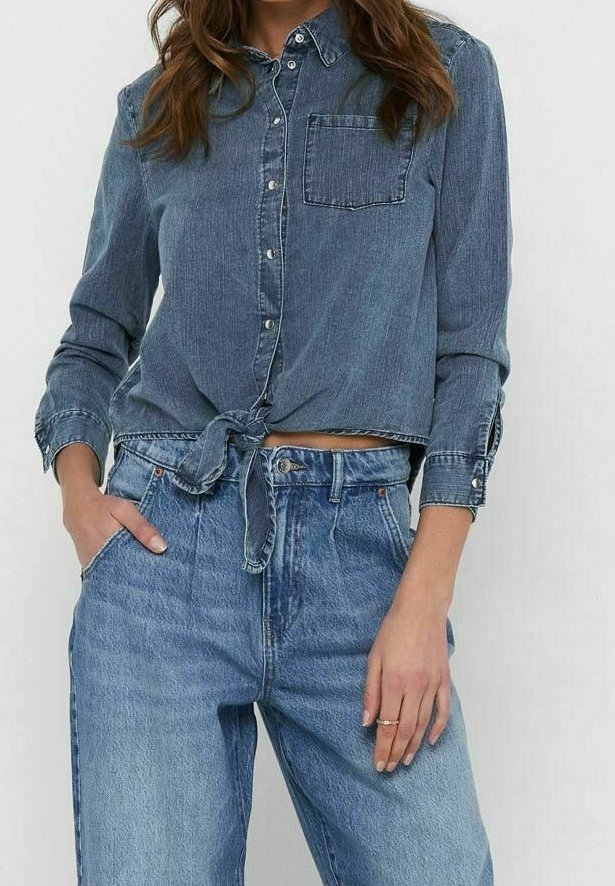 Camicia in denim blu scuro con chiusura a bottoni, annodata in vita, dotata di taschino sul petto e colletto classico. Abbinata a jeans a vita alta.