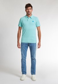 Světle tyrkysové polo tričko, krátké rukávy, dvoubotový knoflíkový zapínání, malá modrá nášivka na hrudi. Kombinováno se světle modrými džíny a bílými teniskami.