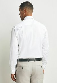 Weißes Langarmhemd mit geknöpften Manschetten, von hinten betrachtet. Kombiniert mit hellgrauen Hosen und einem schwarzen strukturierten Gürtel. Einfaches Design.