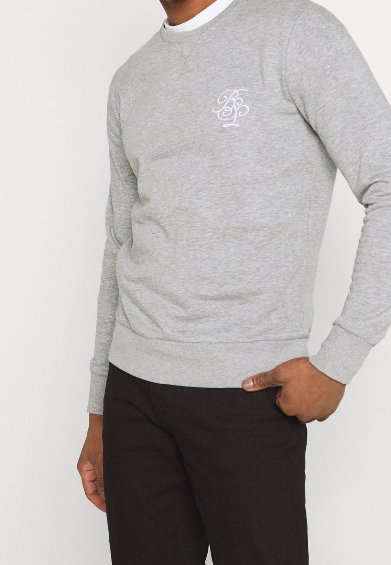 Homme portant un sweat-shirt gris clair à manches longues avec un logo blanc sur la poitrine et un pantalon noir, main dans la poche sur fond uni.