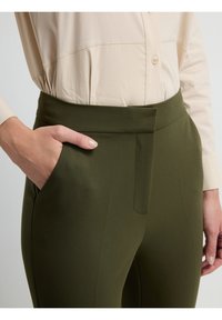Olijfgroene op maat gemaakte broek met een brede tailleband, een vlakke voorkant en zijzakken. De stof lijkt glad en gestructureerd.