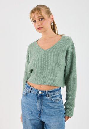 Maglione verde a maglia, con scollo a V, texture a coste e maniche lunghe, abbinato a jeans in denim blu a vita alta.