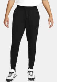 Pantaloni sportivi neri con vita regolabile, design aderente e logo sulla coscia sinistra. Abbinati a sneakers bianche con dettagli neri.