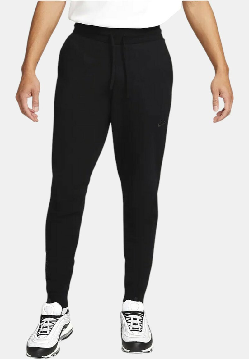 Pantaloni sportivi neri con vita regolabile, design aderente e logo sulla coscia sinistra. Abbinati a sneakers bianche con dettagli neri.