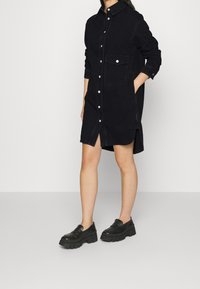 Svart denimklänning med knäppning fram, krage, långa ärmar, sidorfickor och rundad nederkant. Bärs med kraftiga svarta skor.