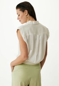 Blusa bianca senza maniche con colletto, texture morbida e un sottile dettaglio plissettato sul retro, abbinata a pantaloni verde chiaro.