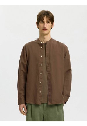 Mladý muž stojaci pred jednoduchým pozadím, oblečený v hnedej košeli na gombíky cez hnedé tričko a zelené nohavice.