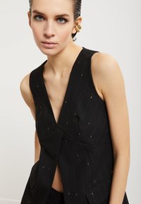 Czarny bezrękawnik z dekoltem w kształcie V, zdobiony złotymi prążkami, o gładkiej fakturze i z węzłowym detalem z przodu.