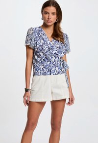 Blauw en witte bloemen wikkel-top met korte mouwen, V-hals, strik in de taille, gecombineerd met witte shorts. Lichte stof, ingewikkeld patroon.