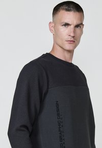 Sudadera negra con parte superior de punto texturizado y panel inferior liso. Presenta un texto impreso en negrita en el costado y un diseño de cuello redondo.