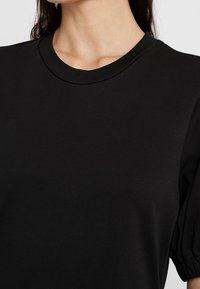 Gros plan d'une personne portant un haut noir uni à col rond avec des manches courtes bouffantes et des cheveux raides et foncés.