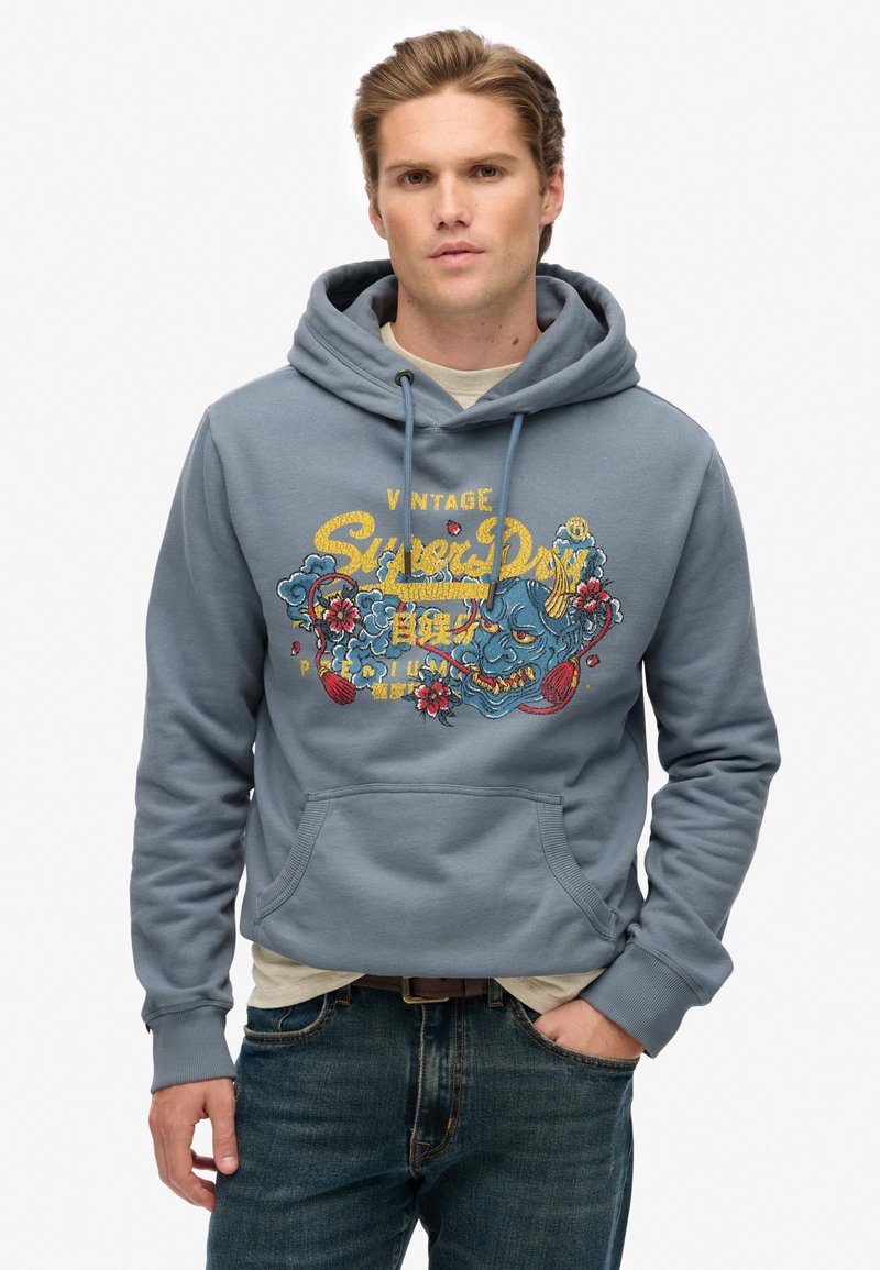 Sudadera con capucha azul y bolsillo frontal, con un estampado gráfico colorido y texto. Fabricada con material suave, presenta puños acanalados.
