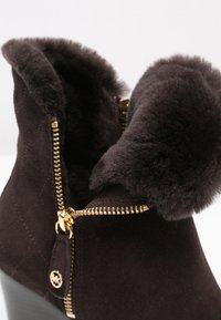 Mörkbruna ankelboots i mocka med en fuskpälskrage, guldfärgade dragkedjor och ett quiltat mönster på sidan. Klacken är en robust blockdesign.