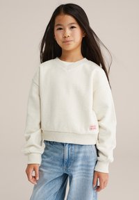 Felpa bianca con macchie, scollo a V, polsini a costine e un'etichetta ricamata rosa che dice "PICCOLE COSE BELLE STANNO ARRIVANDO." Abbinata a jeans blu.