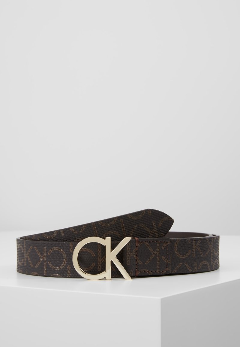 Ceinture marron foncé avec motif répétitif "CK" et grande boucle logo "CK" dorée posée à plat sur une surface blanche.